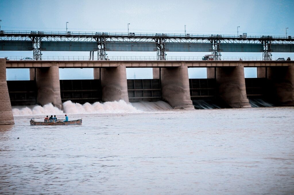 Vista da barragem de uma usina hidrelétrica de médio porte. No rio, é possível ver um bote tripulado por quatro pessoas.